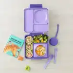 Estuche de silicona OmiePod morado con cubiertos para niños colgado de una lonchera OmieBox